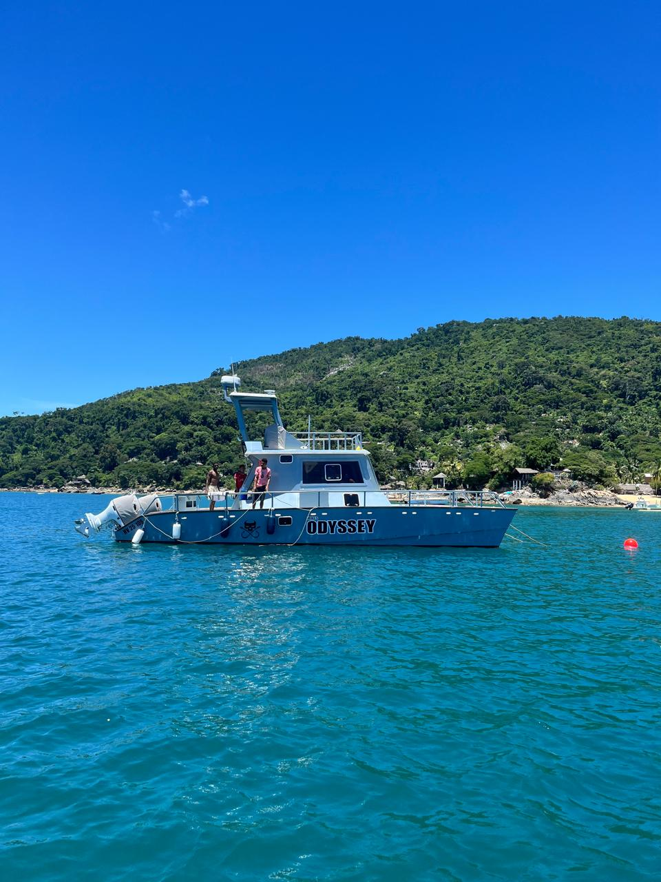 The ODYSSEY sport fishing boat anchored in turquoise water in front of a lush green forested island under a clear blue sky