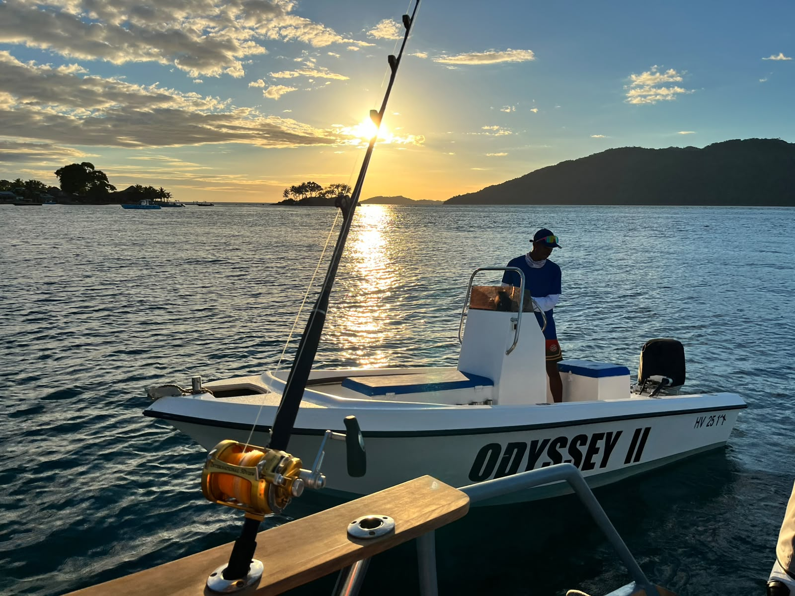 A center-console charter boat named Odyssey II with its captain at the helm at golden hour, an island silhouette on the horizon