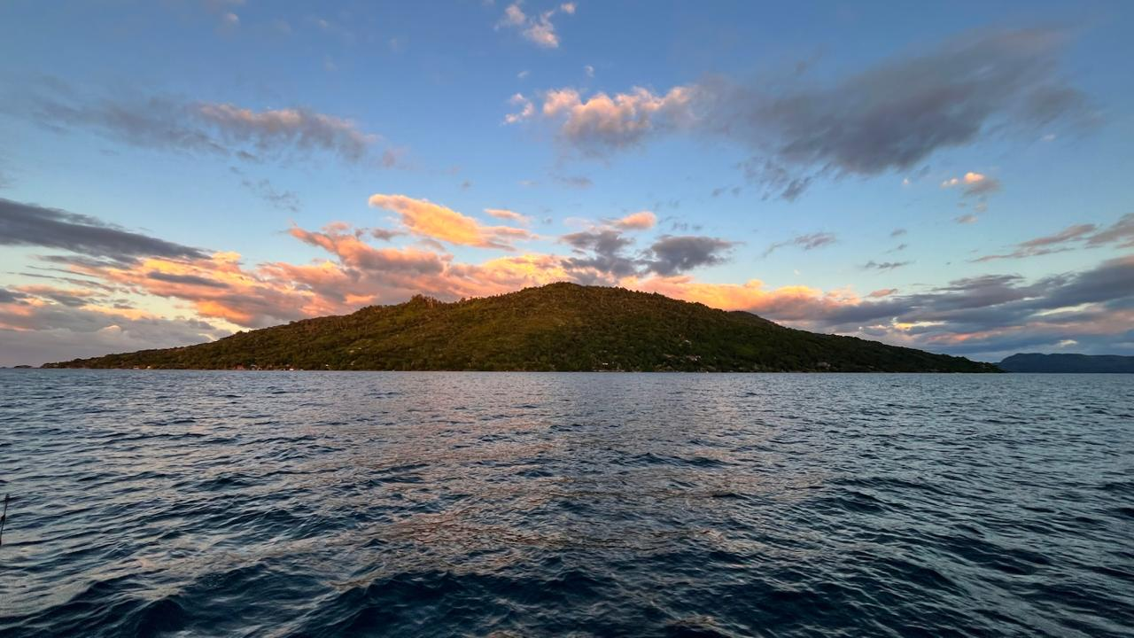 A green forested island at sunset with soft orange clouds over a blue sky, seen across dark rippling water from a boat