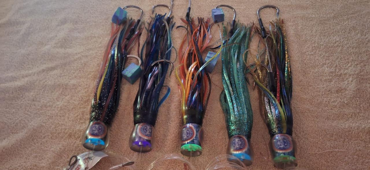 A row of five colourful big-game trolling lures with glittering skirts and hooks laid out on a sandy towel